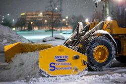 Yellow snow plow with Arctic Sectional Snow-Pusher attachment clearing snow from urban street at night