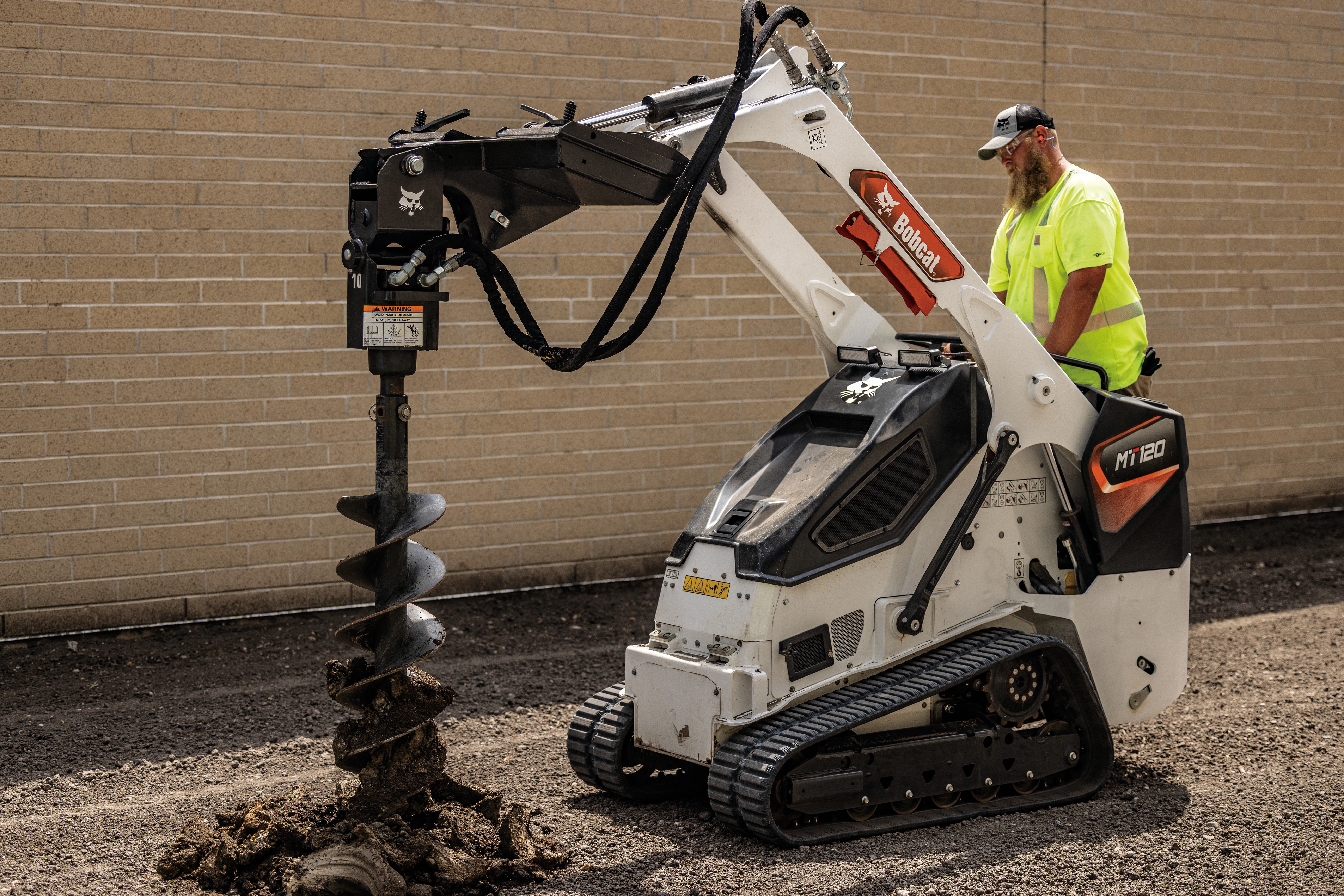 Worker operating white Bobcat mini track loader with auger drill attachment against brick wall