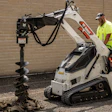 Worker operating white Bobcat mini track loader with auger drill attachment against brick wall