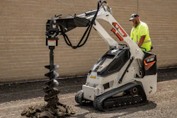 Worker operating white Bobcat mini track loader with auger drill attachment against brick wall