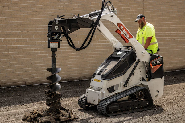 Worker operating white Bobcat mini track loader with auger drill attachment against brick wall