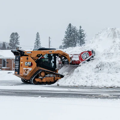 Orange CAT snow removal equipment clearing large piles of snow on a winter road with snow-covered trees in background