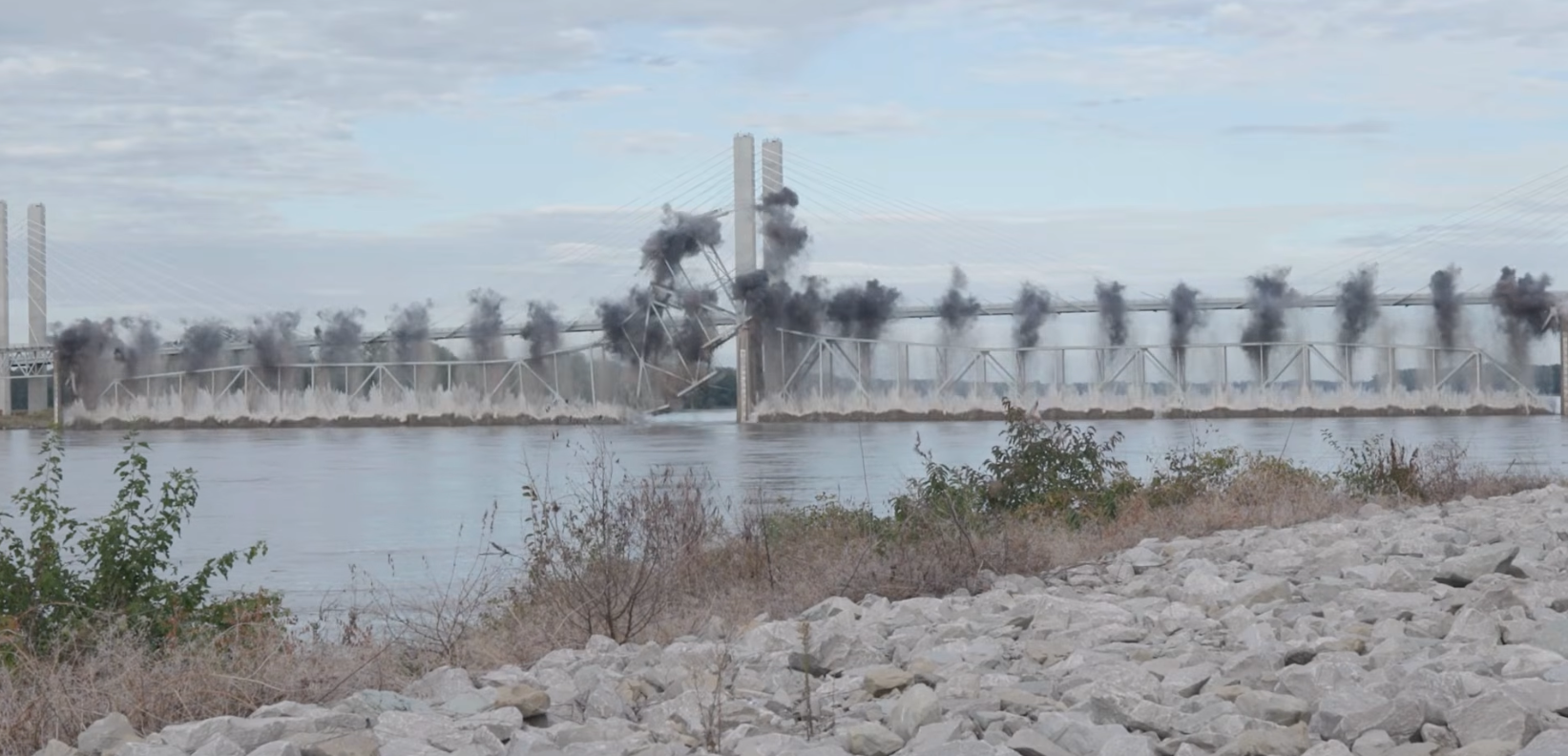 Industrial waterfront with bridge spanning river and factory smokestacks emitting smoke, rocky shoreline in foreground