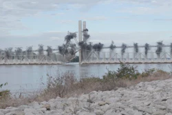 Industrial waterfront with bridge spanning river and factory smokestacks emitting smoke, rocky shoreline in foreground
