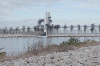 Industrial waterfront with bridge spanning river and factory smokestacks emitting smoke, rocky shoreline in foreground
