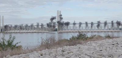 Industrial waterfront with bridge spanning river and factory smokestacks emitting smoke, rocky shoreline in foreground