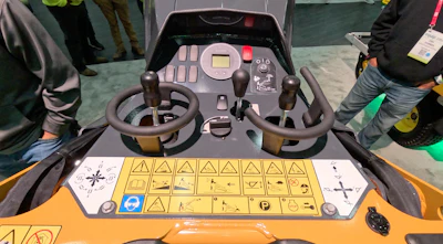 Heavy equipment control console with dual joysticks and yellow button panel displayed at an exhibition.