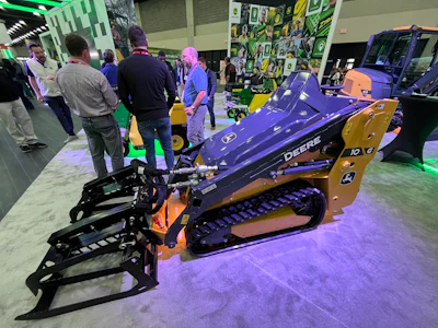 Purple and gold John Deere compact track loader on display at an indoor equipment exhibition with visitors