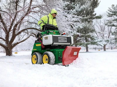 Worker operating green John Deere snow plow with Western brand equipment clearing snow from park pathway in winter