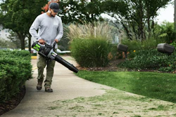 Man using a leaf blower to clear debris from a residential driveway surrounded by landscaping and trees