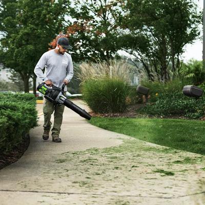 Man using a leaf blower to clear debris from a residential driveway surrounded by landscaping and trees
