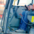 Construction equipment operator in orange safety vest operating machinery from cabin with yellow safety badge visible