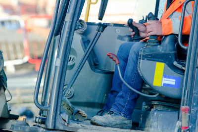 Construction equipment operator in orange safety vest operating machinery from cabin with yellow safety badge visible