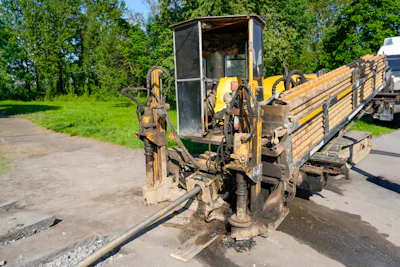 Yellow and black horizontal directional drill on asphalt road with wooden trailer and green trees in background