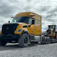 Gold Volvo semi-truck with black grille towing a yellow construction vehicle on gravel site under cloudy sky