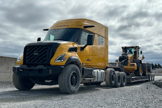 Gold Volvo semi-truck with black grille towing a yellow construction vehicle on gravel site under cloudy sky