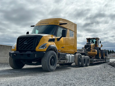 Gold Volvo semi-truck with black grille towing a yellow construction vehicle on gravel site under cloudy sky