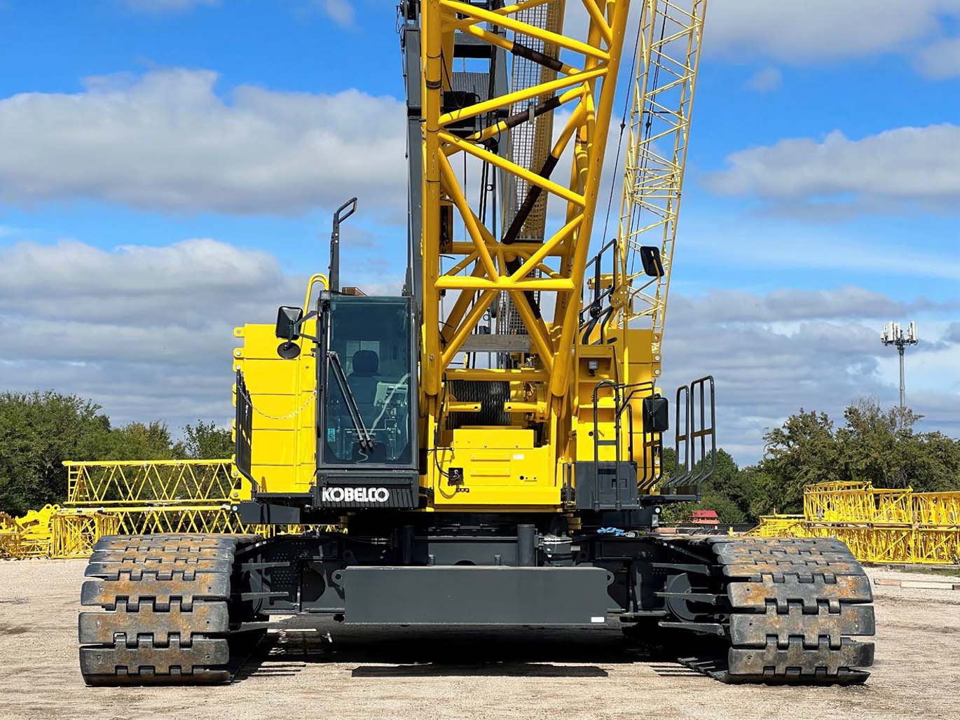 Yellow and black Kobelco crawler crane with lattice boom positioned on gravel ground against blue sky