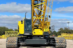 Yellow and black Kobelco crawler crane with lattice boom positioned on gravel ground against blue sky