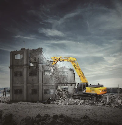 Yellow excavator demolishing abandoned concrete building under stormy sky
