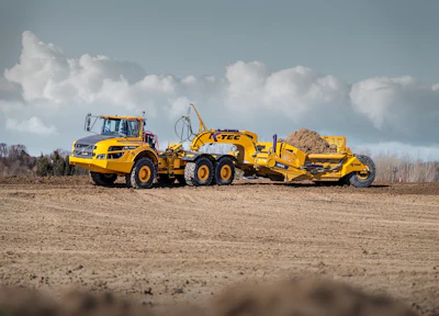 Yellow K-Tec 1232 ADT scraper being pulled by Komatsu articulated dump truck chassis on a dirt site under blue sky