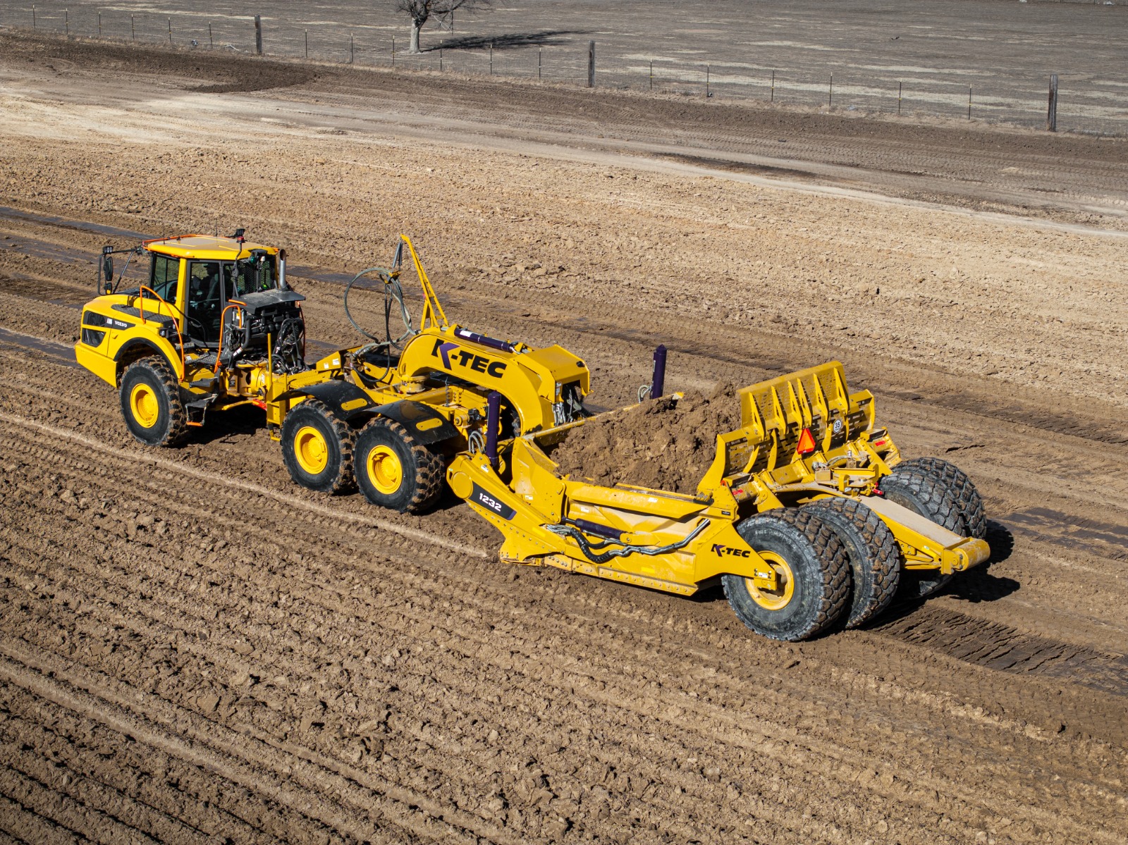 Yellow K-Tec 1232 ADT scraper being pulled by Komatsu articulated dump truck chassis on unpaved construction site