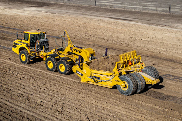 Yellow K-Tec 1232 ADT scraper being pulled by Komatsu articulated dump truck chassis on unpaved construction site