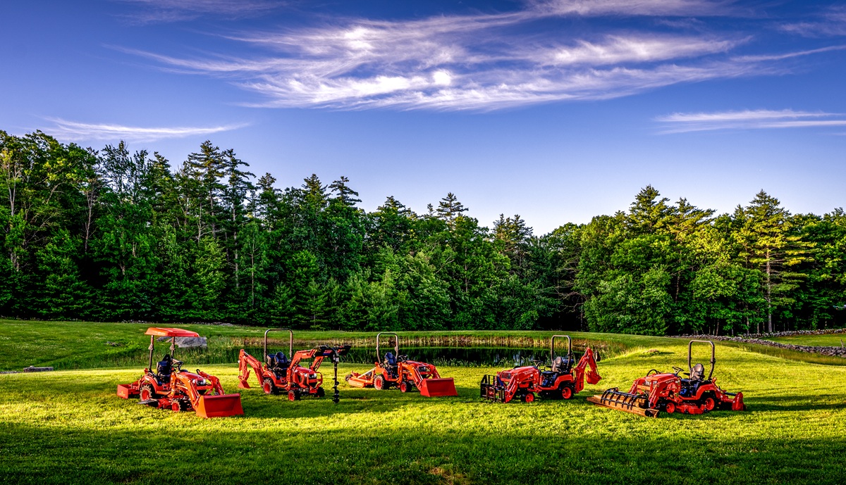 Red farm equipment and machinery lined up in a sunny green field with forest backdrop