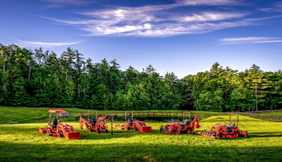 Red farm equipment and machinery lined up in a sunny green field with forest backdrop