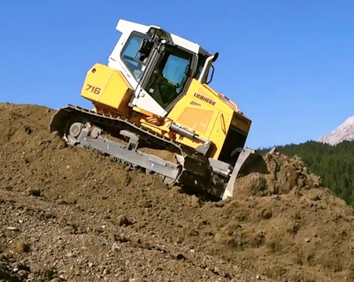 Yellow and white Liebherr 716 tracked bulldozer operating on rocky terrain with forested mountains in background