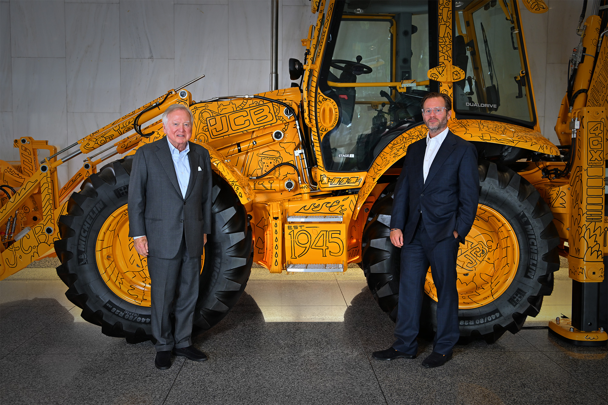 Two businessmen in suits stand in front of a yellow JCB telescopic handler in a showroom setting