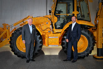 Two businessmen in suits stand in front of a yellow JCB telescopic handler in a showroom setting