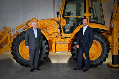 Two businessmen in suits stand in front of a yellow JCB telescopic handler in a showroom setting