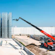 Red telehandler boom lift operating at industrial construction site with steel framework and white warehouse building
