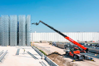 Red telehandler boom lift operating at industrial construction site with steel framework and white warehouse building
