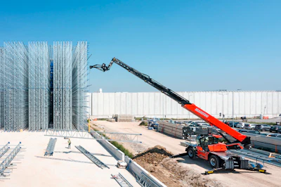 Red telehandler boom lift operating at industrial construction site with steel framework and white warehouse building