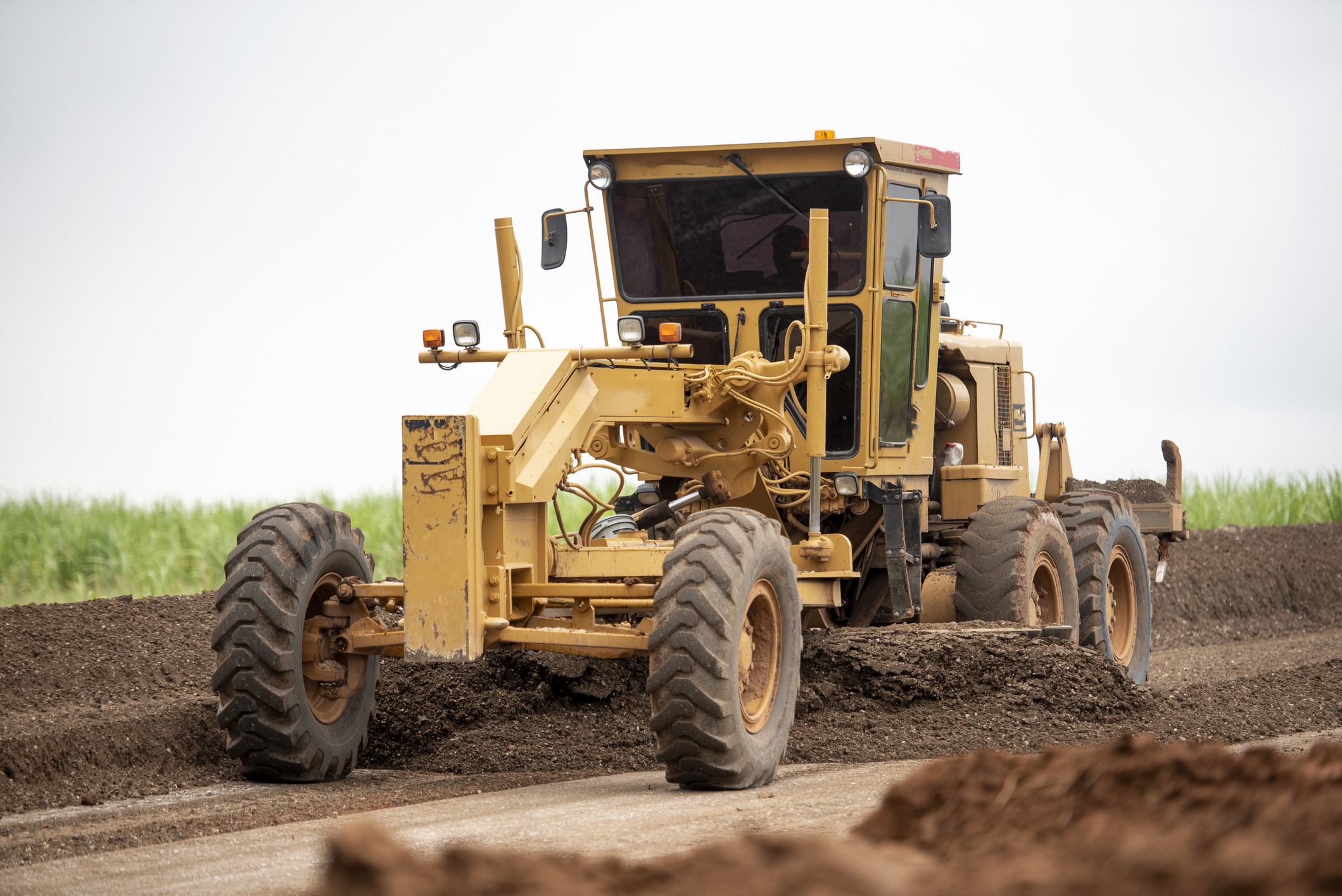 Yellow motor grader leveling dirt on a road construction site with green fields in background