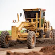 Yellow motor grader leveling dirt on a road construction site with green fields in background