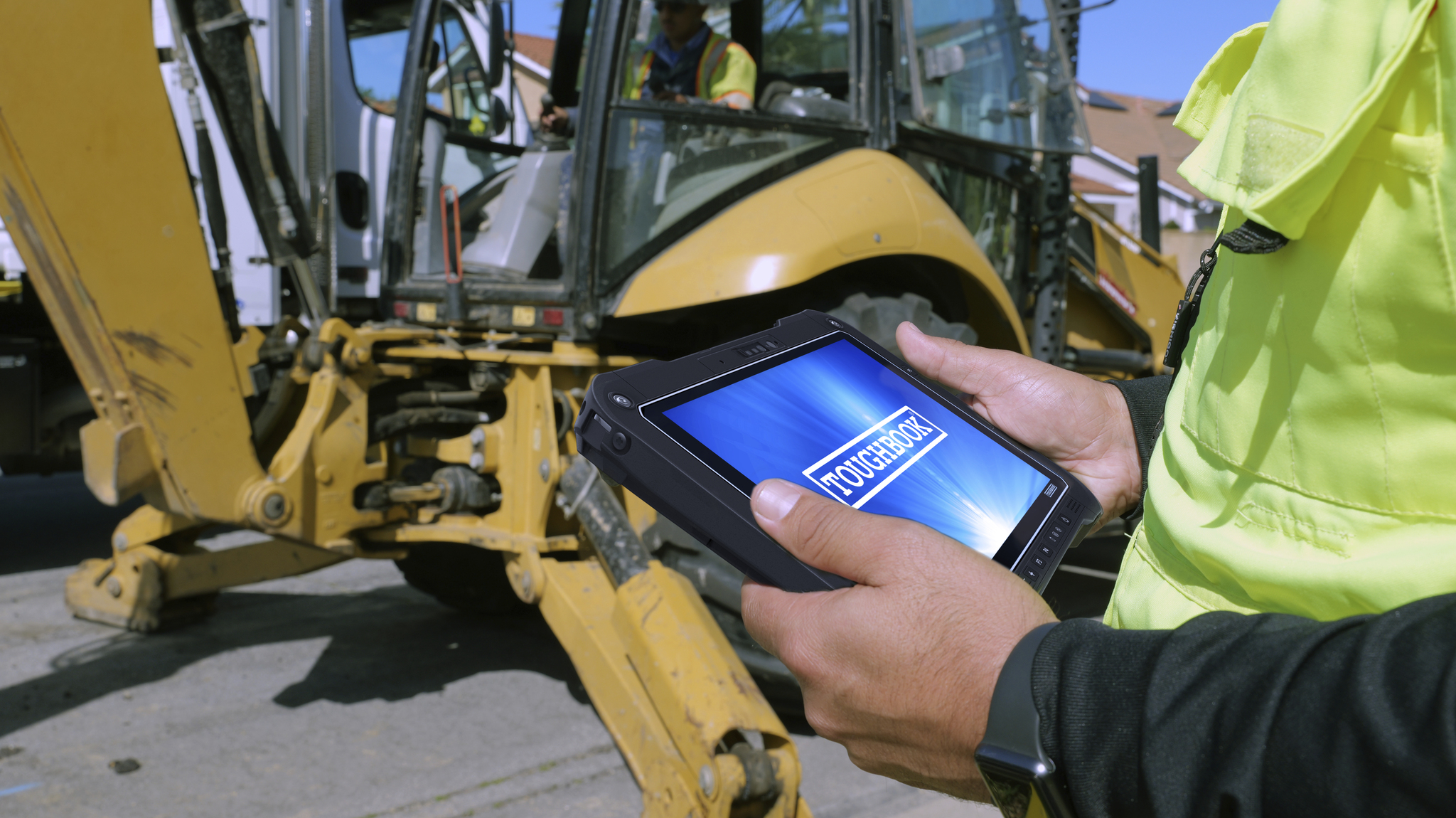 Construction worker holding Panasonic Toughbook G3 tablet with blue screen display near yellow backhoe on jobsite