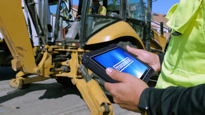 Construction worker holding Panasonic Toughbook G3 tablet with blue screen display near yellow backhoe on jobsite