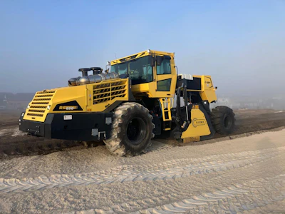 Yellow motor grader construction equipment operating on sandy terrain with city skyline in background
