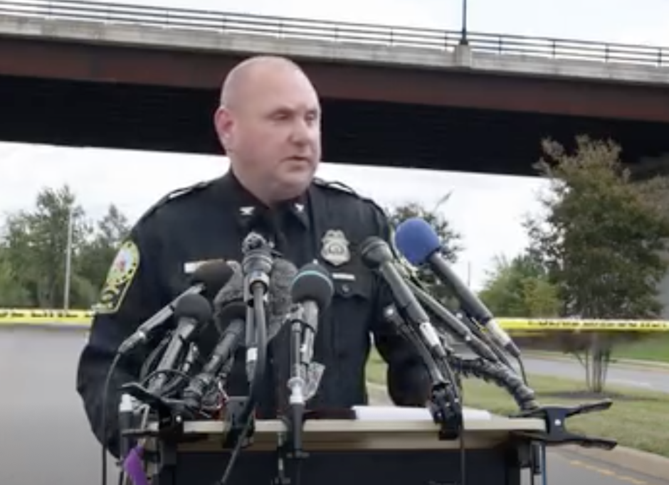 Police officer speaking at press conference with multiple microphones and yellow crime scene tape visible
