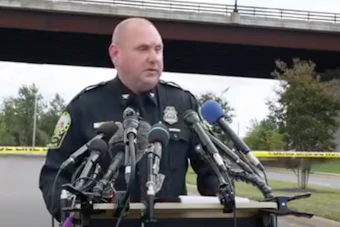 Police officer speaking at press conference with multiple microphones and yellow crime scene tape visible