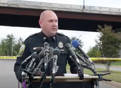 Police officer speaking at press conference with multiple microphones and yellow crime scene tape visible