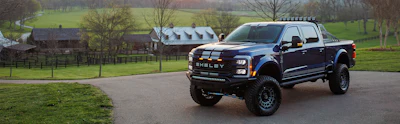 Custom Shelby pickup truck parked on driveway with rural farmland and buildings in background