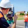 Surveyor in safety gear using theodolite to measure land with assistant in background