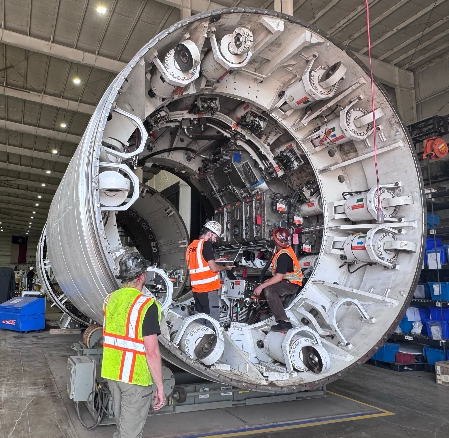 Technicians in safety gear inspect a large cylindrical spacecraft module with docking ports in an industrial hangar facility.