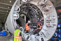 Technicians in safety gear inspect a large cylindrical spacecraft module with docking ports in an industrial hangar facility.