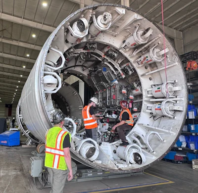 Technicians in safety gear inspect a large cylindrical spacecraft module with docking ports in an industrial hangar facility.
