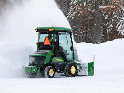 Green John Deere snow plow clearing snow on a winter road lined with snow-covered pine trees